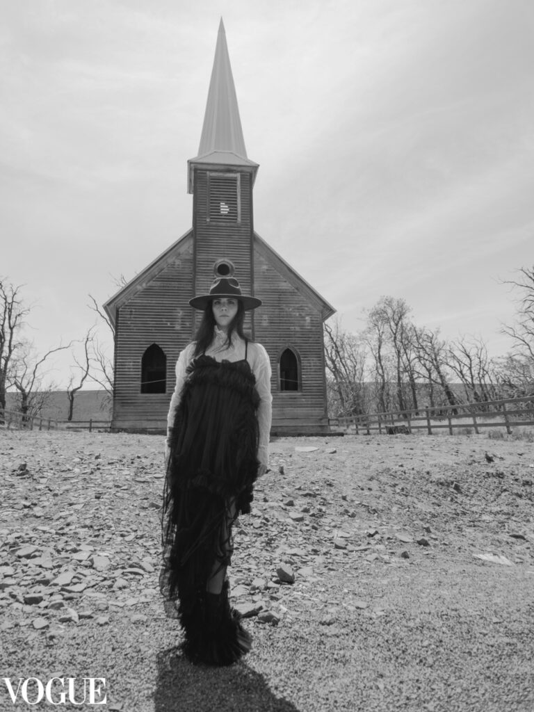 Black and white photo published in Vogue. Portrait of a woman dressed in black, standing in front of an abandoned church. Photo en noir et blanc publiée dans Vogue. Une femme vêtue de noir, se tient devant une église abandonnée.