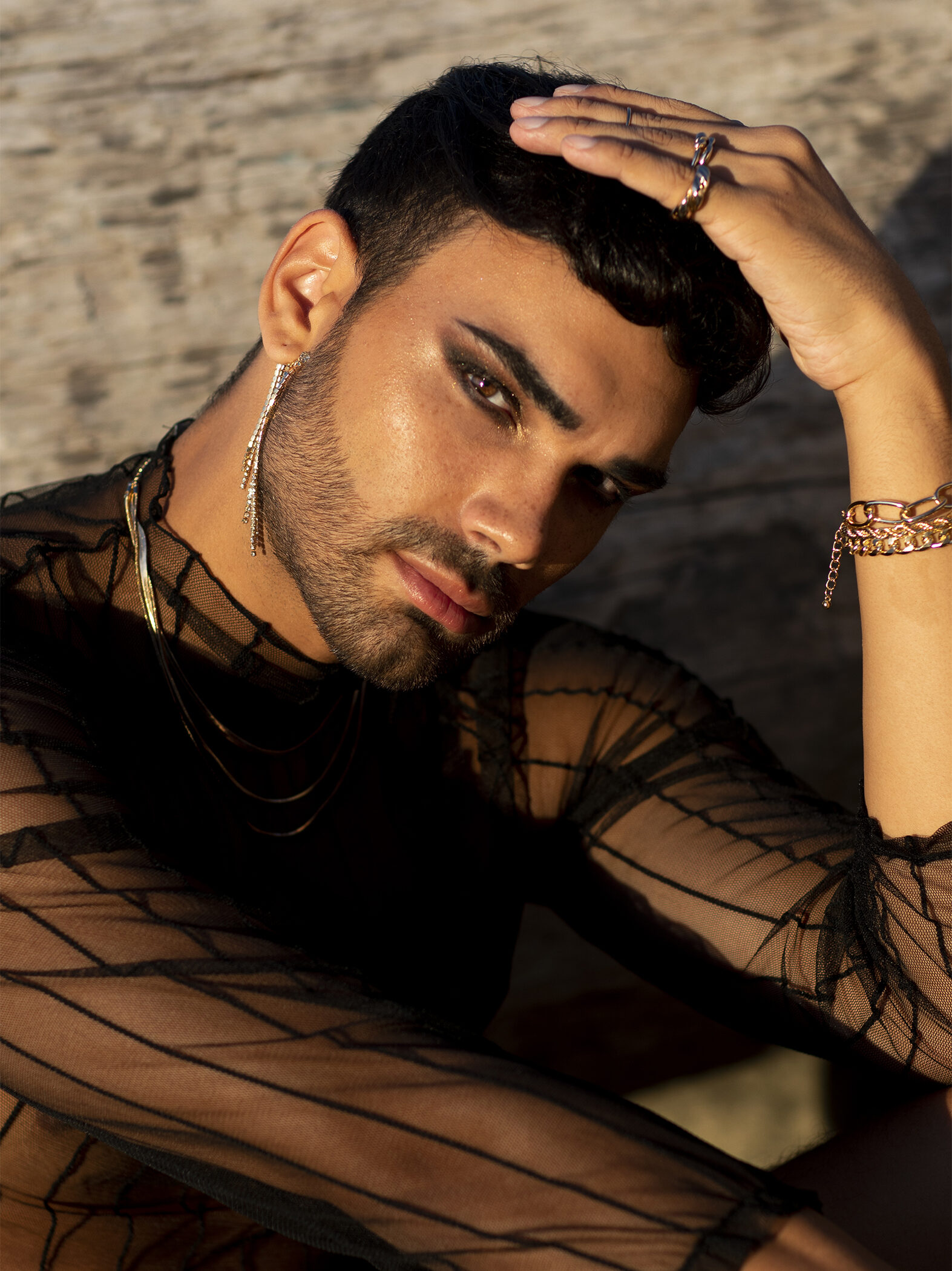 Beauty fashion photography. A beautiful native man wearing jewelry and smoky makeup, sitting at the beach during golden hour. Portrait de photo de mode. Un homme portant des bijoux et un maquillage feutré, est assis sur la plage, sous le soleil de fin de journée.